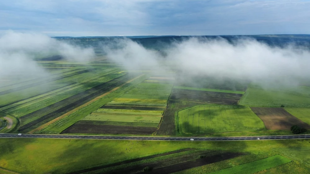 An aerial view of a highway cutting through green and patchy agricultural fields, with a layer of mist and clouds hovering above the landscape.