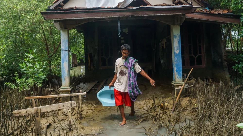 A man in red shorts and a white tshirt carries a light blue toilet commode from a ruined house, walking down a path through a muddy flooded landscape.
