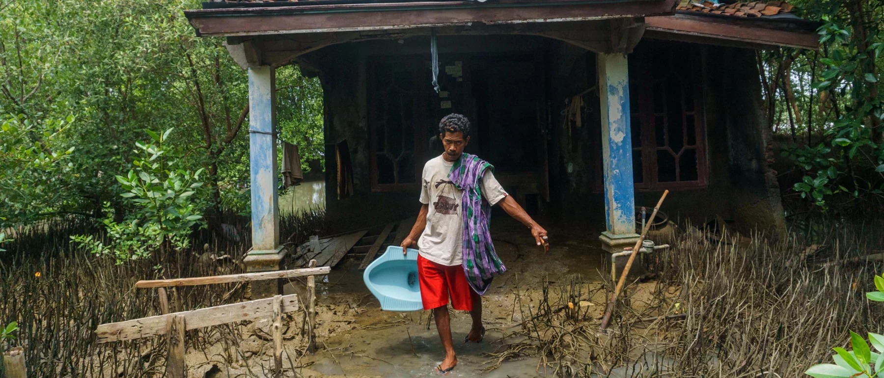 A man in red shorts and a white tshirt carries a light blue toilet commode from a ruined house, walking down a path through a muddy flooded landscape.
