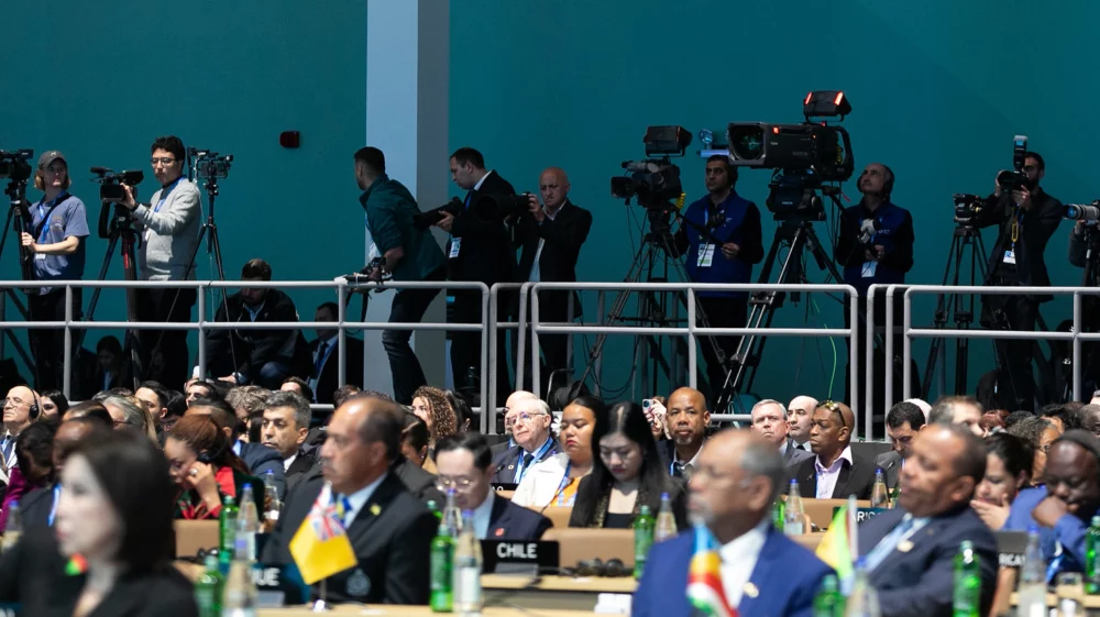A diverse group of delegates seated at an international conference, with country nameplates visible, such as Chile and Canada. Behind the delegates, a raised platform is occupied by camera operators and media personnel capturing the event.