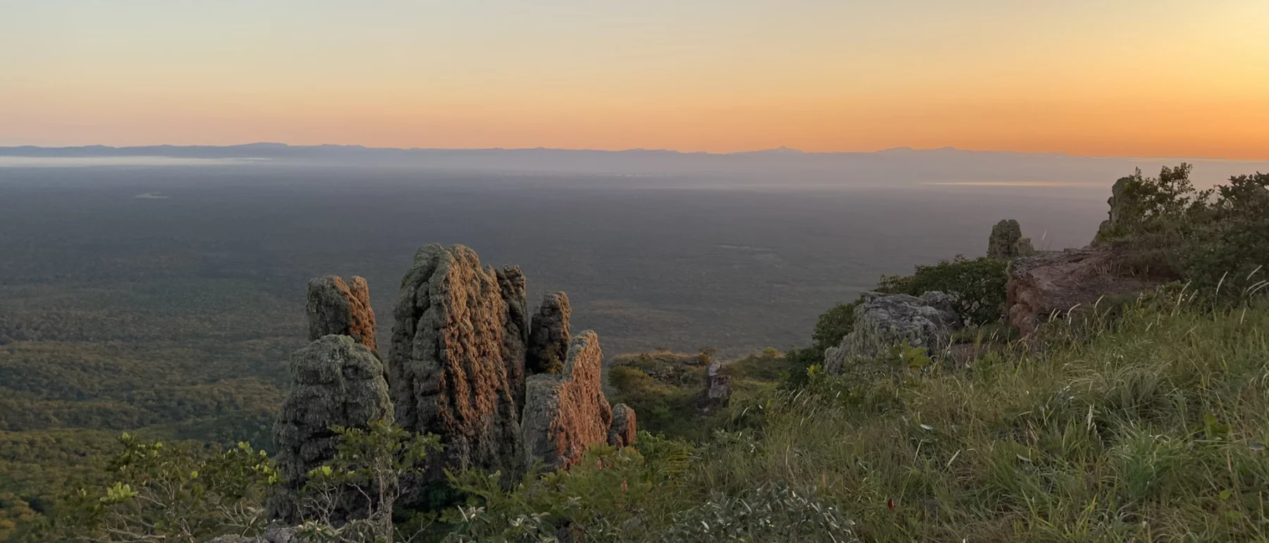 sunset over ocean at the horizon, with outcrops of red rocks and green shrubs in the foreground