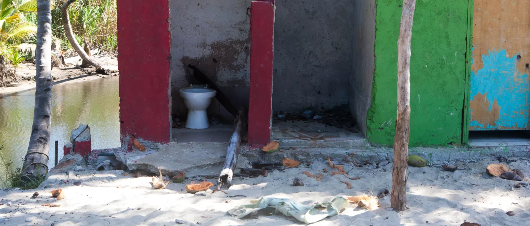 A broken down bathroom in the middle of a beach. A toilet with no door. Colored walls and palm trees.
