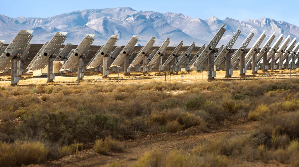 several rows of white metal solar panels viewed from the side, with dry yellow grass and green shrubs in the foreground, and light tan mountains in the background with folding rocks.