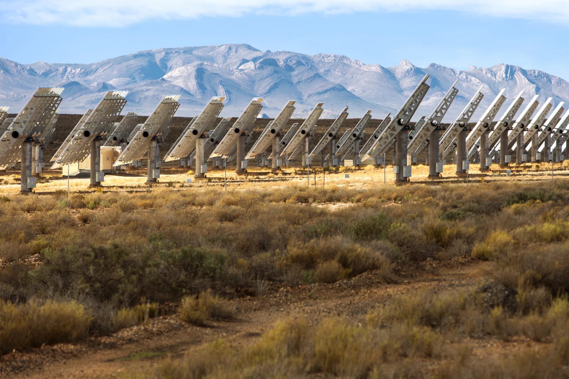 several rows of white metal solar panels viewed from the side, with dry yellow grass and green shrubs in the foreground, and light tan mountains in the background with folding rocks.