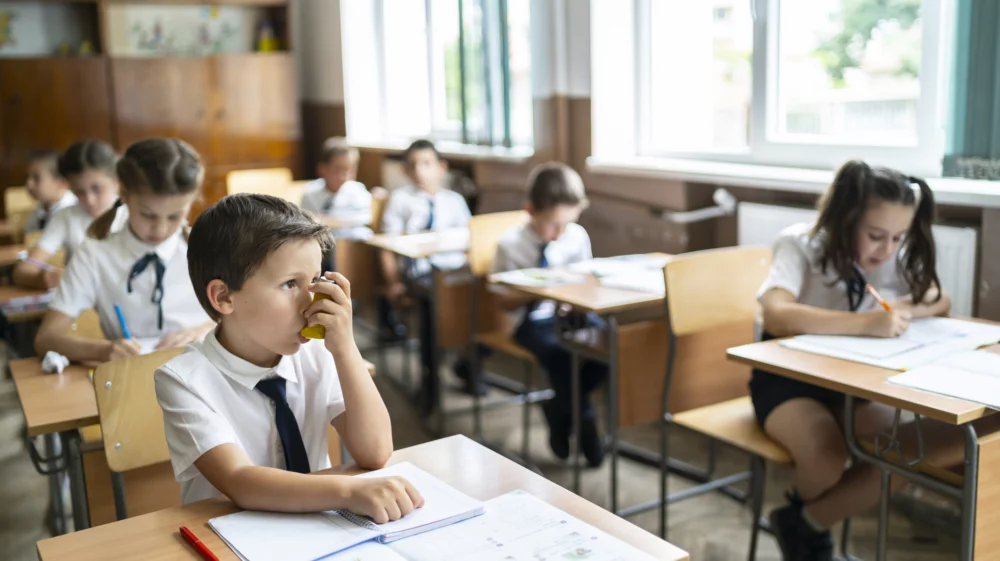 Elementary school boy using asthma pump inhaler at school during lessons.