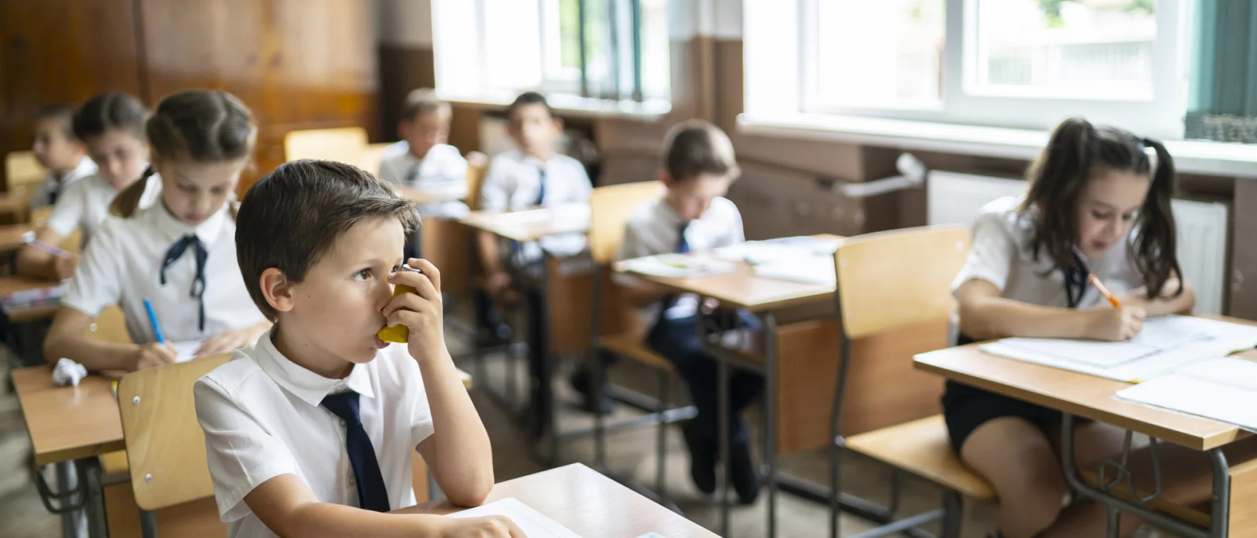 Elementary school boy using asthma pump inhaler at school during lessons.