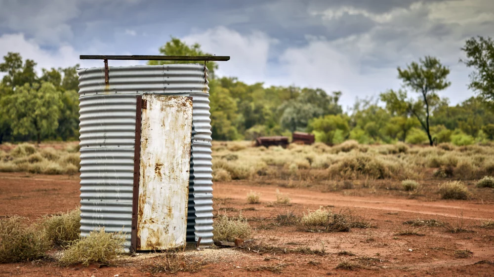 A corrugated metal tube with a hanging rusty metal door stands on flat red dirt with trees in the far distance and a rusty tractor.