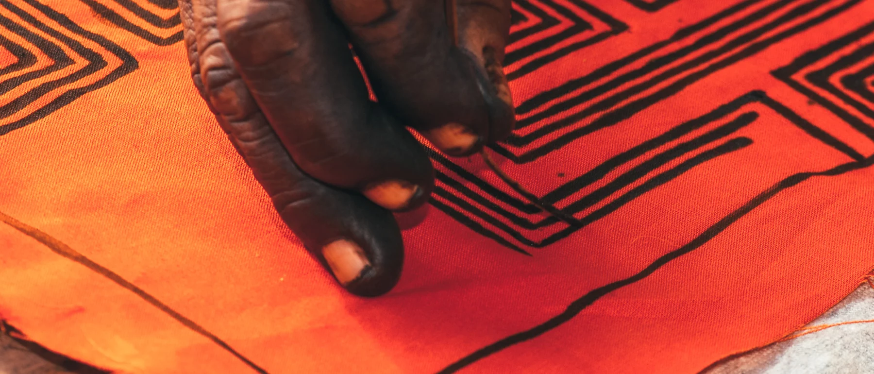 A close-up of an Indigenous person’s hand painting intricate black geometric patterns on a vibrant orange fabric.