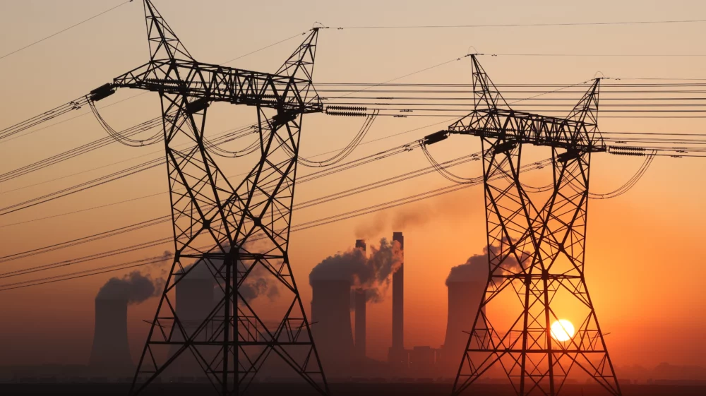 Two electricity towers in the foreground are illuminated by an orange sunset and four nuclear tower stacks with steam clouds and two chimneys with smoke plumes from coal power plant in South Africa.