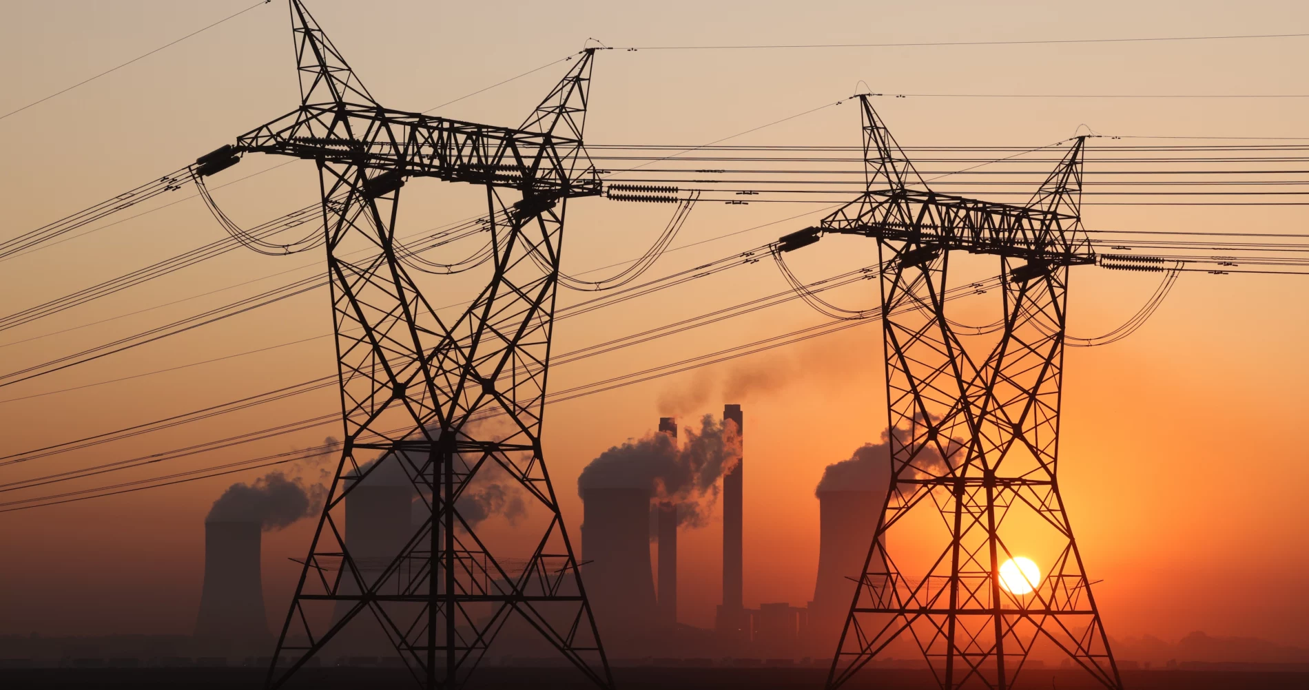 Two electricity towers in the foreground are illuminated by an orange sunset and four nuclear tower stacks with steam clouds and two chimneys with smoke plumes from coal power plant in South Africa.