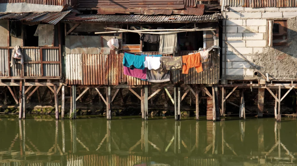 Informal stilt housing made of corrugated metal sheets and concrete blocks extends over a murky, greenish water body. Clothes hang from a line in front of the dwellings.