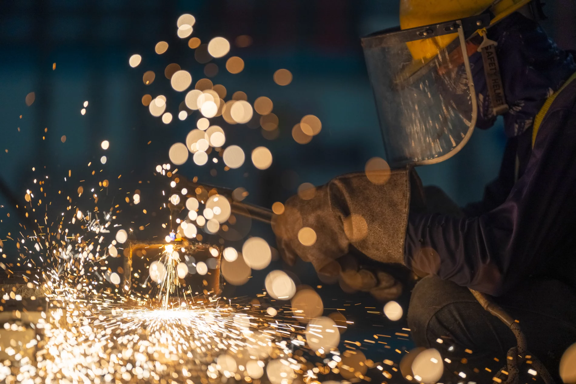 Glowing hot white and gold sparks fly as a worker with a welding mask, heavy gloves and tool leans in to the surface.