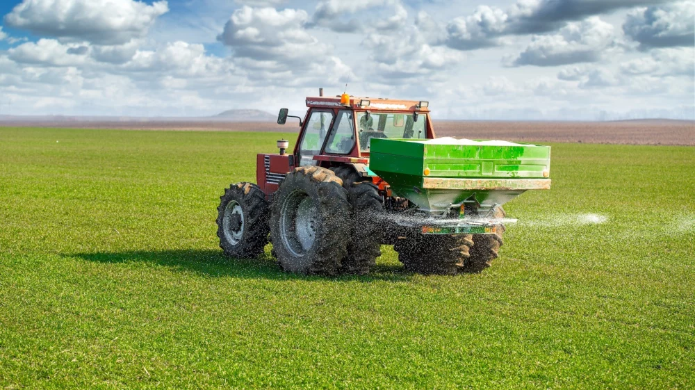 A tractor spreading fertilizer on a green wheat field under a partly cloudy sky. The machine has a green fertilizer spreader attached at the back, and white particles can be seen dispersing across the field.
