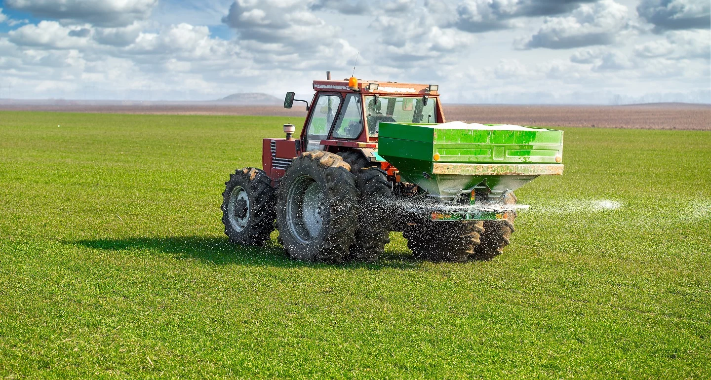 A tractor spreading fertilizer on a green wheat field under a partly cloudy sky. The machine has a green fertilizer spreader attached at the back, and white particles can be seen dispersing across the field.