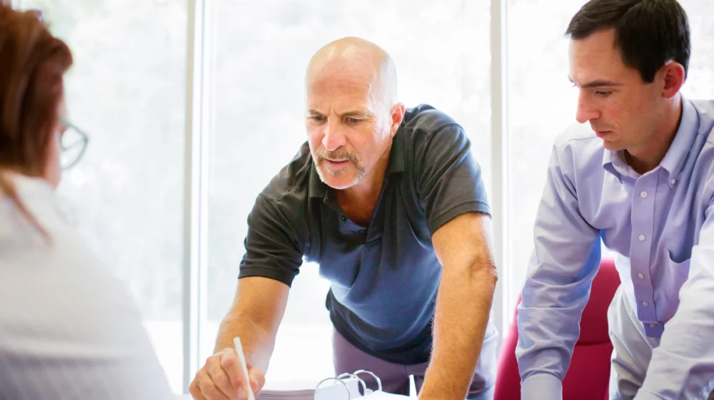 Troubled man pointing to small print on paper in front of unphased bureaucratic person. His friend is by his side.