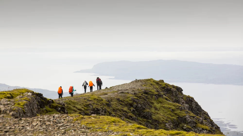 A group of hikers with brightly colored backpacks trek along a rocky, moss-covered ridge, overlooking a vast, misty landscape and distant coastline under a grey sky. The scene captures an expansive, serene wilderness with a sense of adventure.