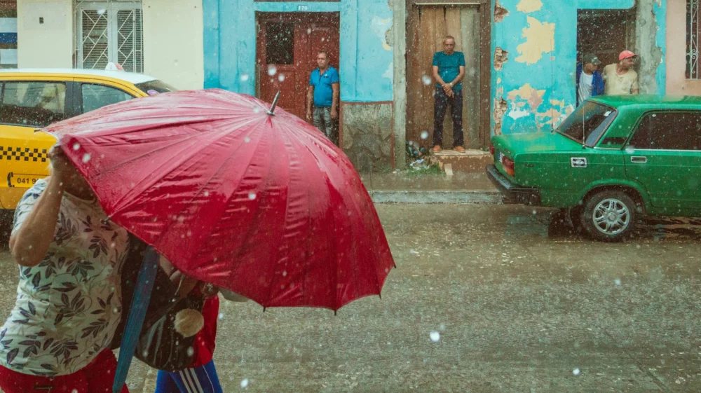 People walking under a large red umbrella during a rain shower on a city street, with colourful, weathered buildings in the background and onlookers standing in doorways.