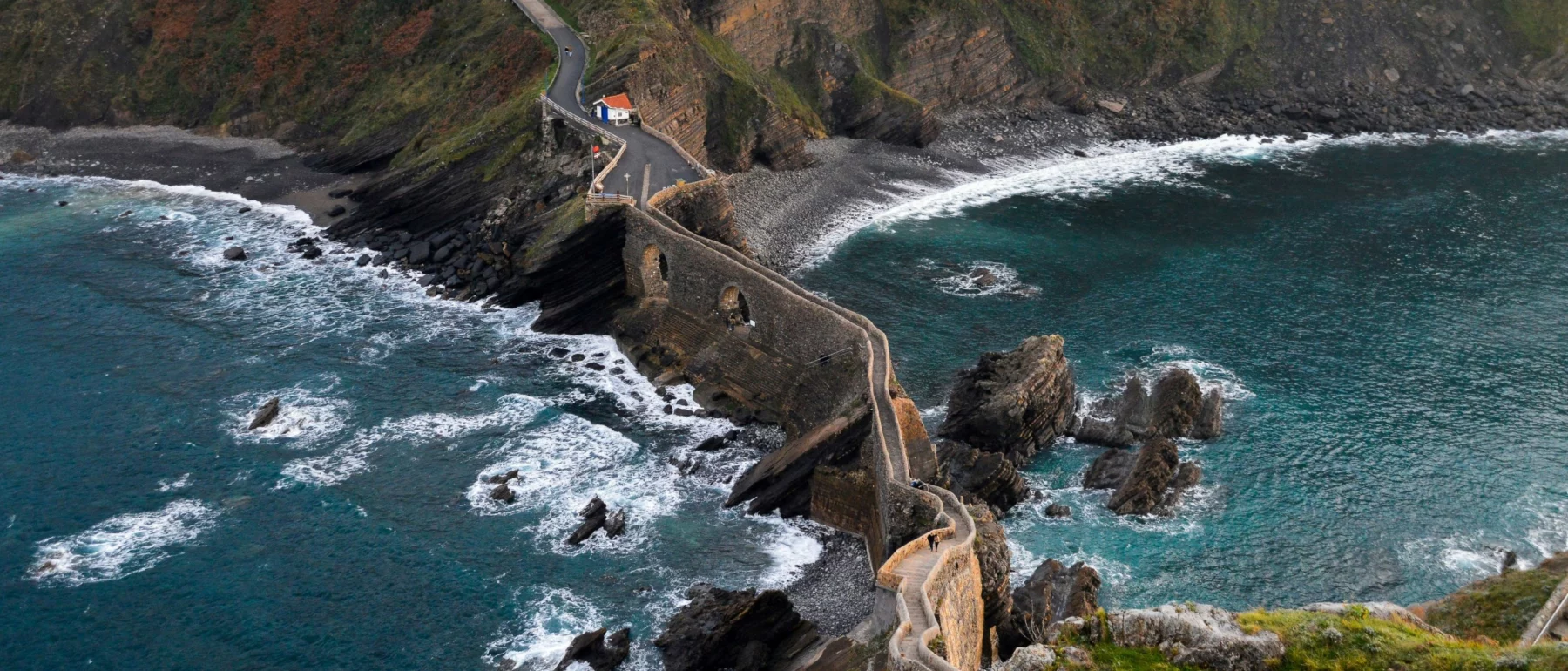A winding stone pathway leading to a small white chapel on top of a rugged, rocky peninsula jutting into the blue ocean, surrounded by steep cliffs and crashing waves.