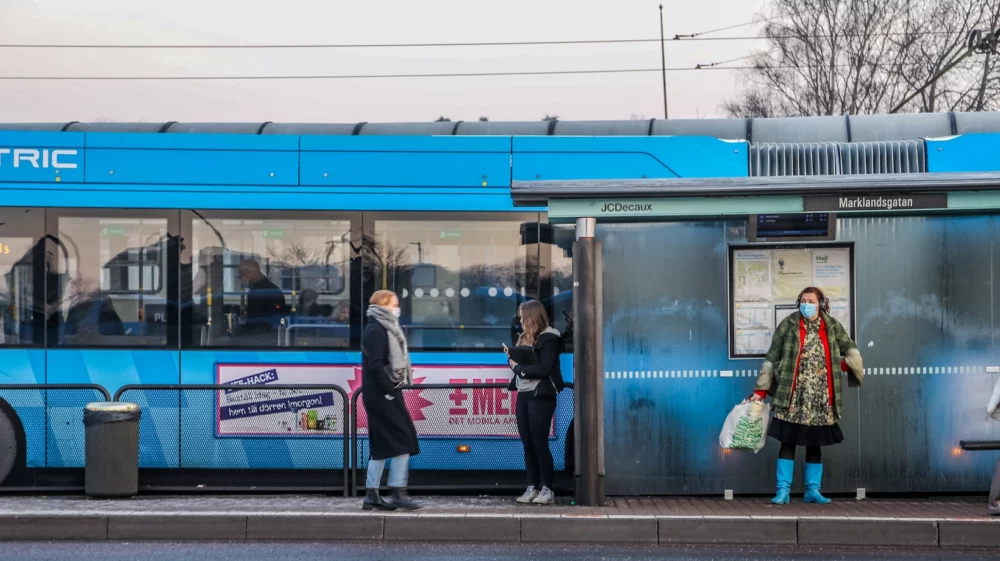 A blue electric bus at a bus stop named Marklandsgatan in Sweden. Passengers, some wearing face masks, wait and board the bus. The bus display shows “Departs in 50 seconds.” The scene reflects a cold day, with people dressed warmly.