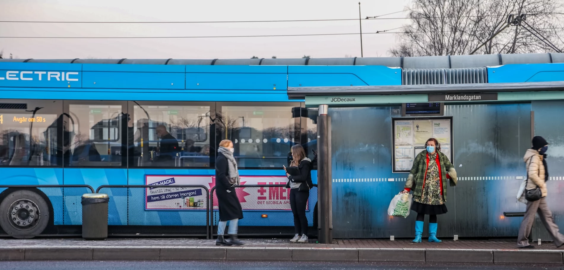 A blue electric bus at a bus stop named Marklandsgatan in Sweden. Passengers, some wearing face masks, wait and board the bus. The bus display shows “Departs in 50 seconds.” The scene reflects a cold day, with people dressed warmly.