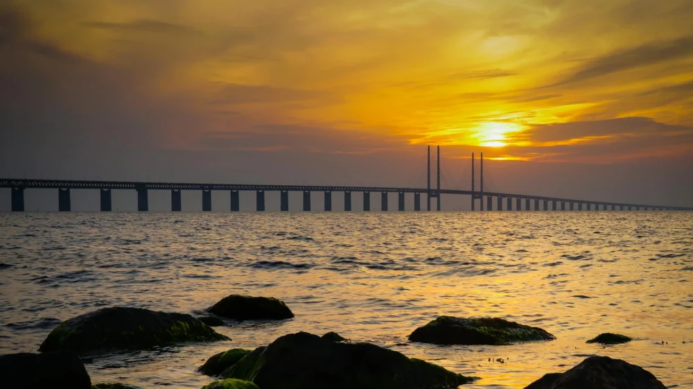 A scenic view of the Öresund Bridge connecting Sweden and Denmark, photographed at sunset over calm waters with rocky foreground.