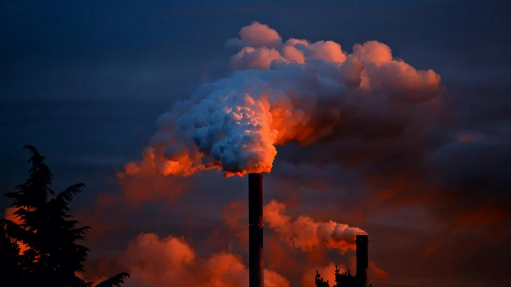 A tall industrial smokestack releasing thick clouds of smoke into a darkening sky. The smoke is illuminated by a warm, reddish-orange glow, contrasting with the blue tones of the surrounding sky.