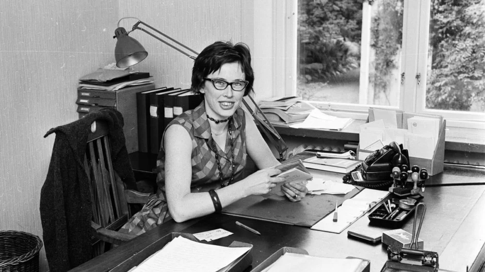 A woman sitting at a desk in an office, smiling at the camera while holding a document. The desk is cluttered with papers, office supplies and a rotary telephone. The room has a large window in the background, letting in natural light. The image is from the 1960s.