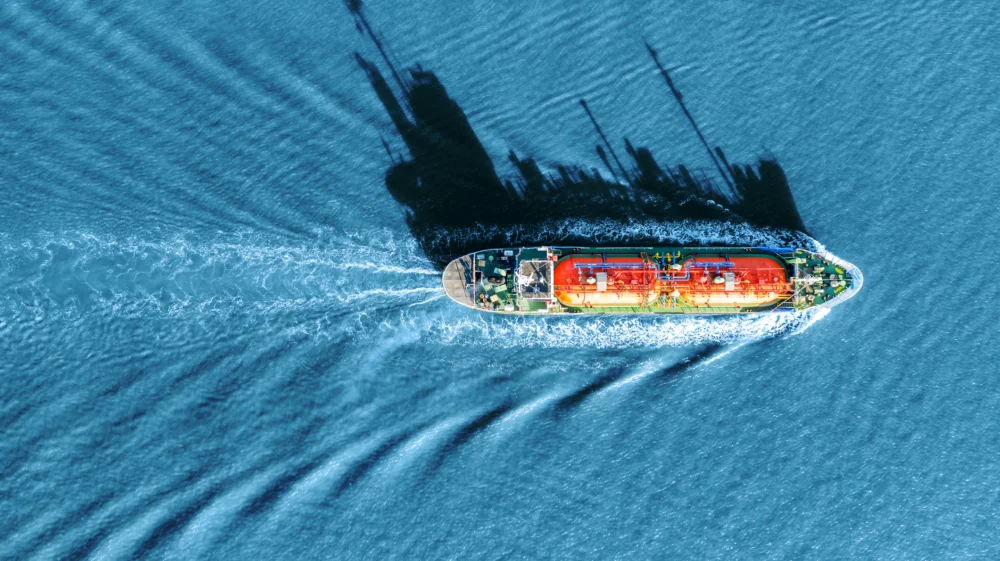 aerial view of an orange-and-green ship tanker on a bright blue sea, with black shadow projected to the top of the image of towers and pipes.