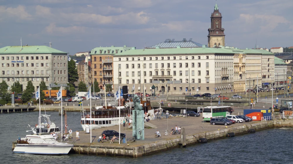 Boats of different sizes are docked at a large cement platform on blue water, with statue and flags, cars and a bus, people sunning themselves in the summer sun on benches, with large buildings in the background.
