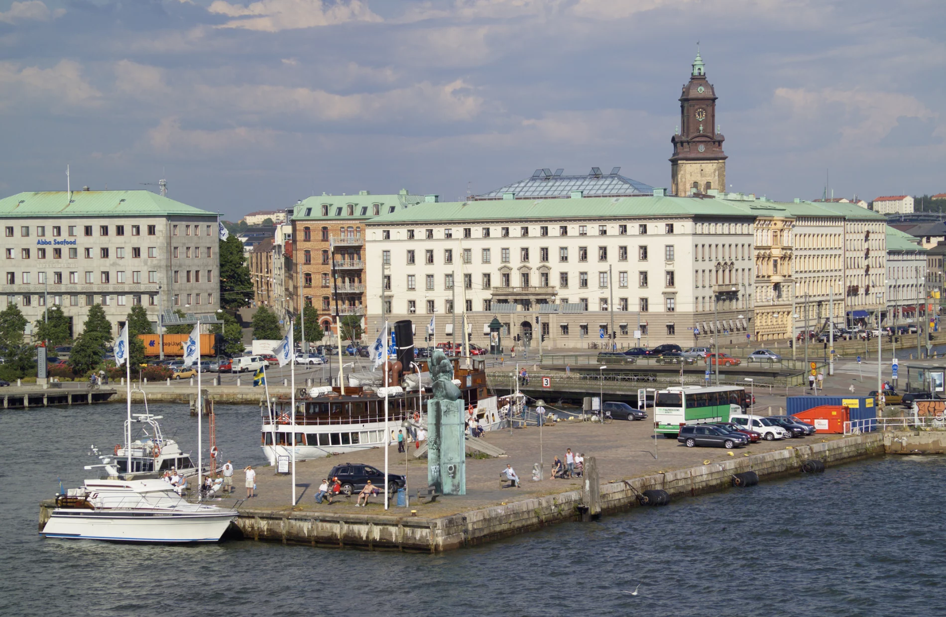 Boats of different sizes are docked at a large cement platform on blue water, with statue and flags, cars and a bus, people sunning themselves in the summer sun on benches, with large buildings in the background.