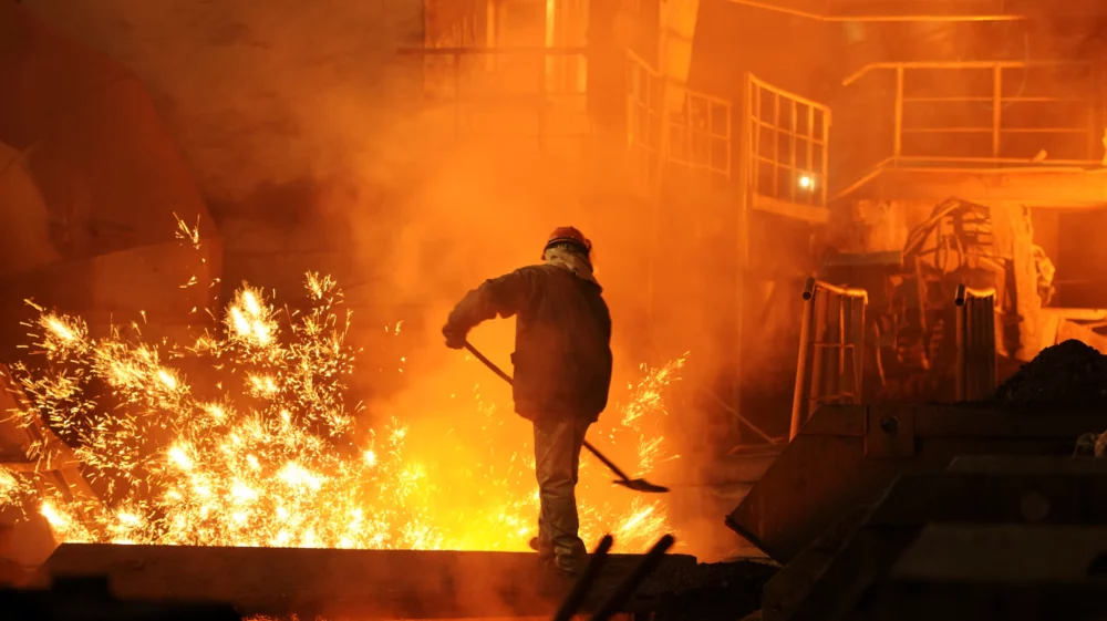 A worker in protective gear operates near molten metal in a brightly lit steelmaking factory, with sparks flying and an industrial backdrop illuminated by an orange glow.