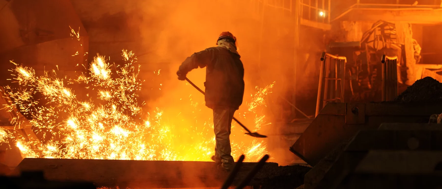 A worker in protective gear operates near molten metal in a brightly lit steelmaking factory, with sparks flying and an industrial backdrop illuminated by an orange glow.