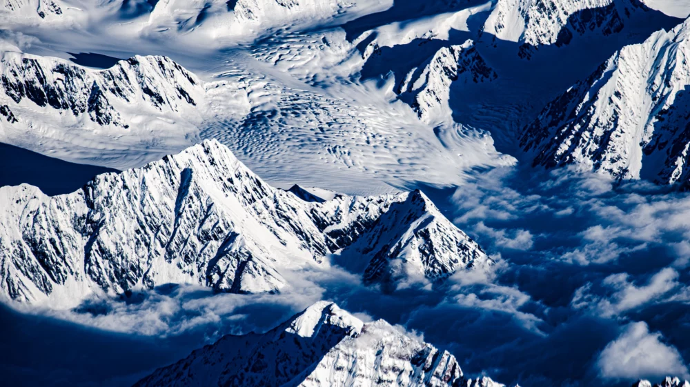 White snowy peaks around a white snow-covered flowing and lumpy glacier, with blue shadows, clouds.