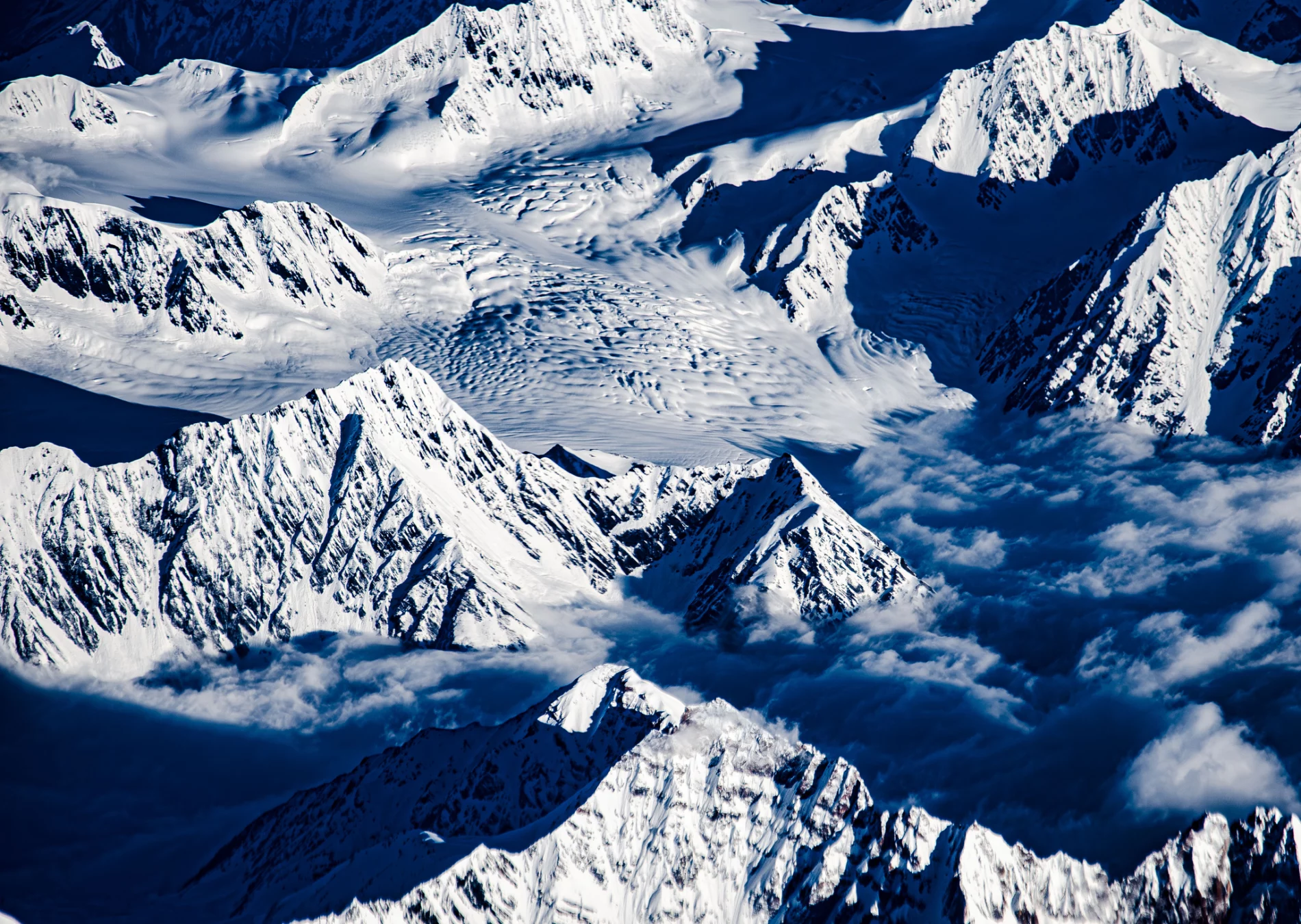 White snowy peaks around a white snow-covered flowing and lumpy glacier, with blue shadows, clouds.