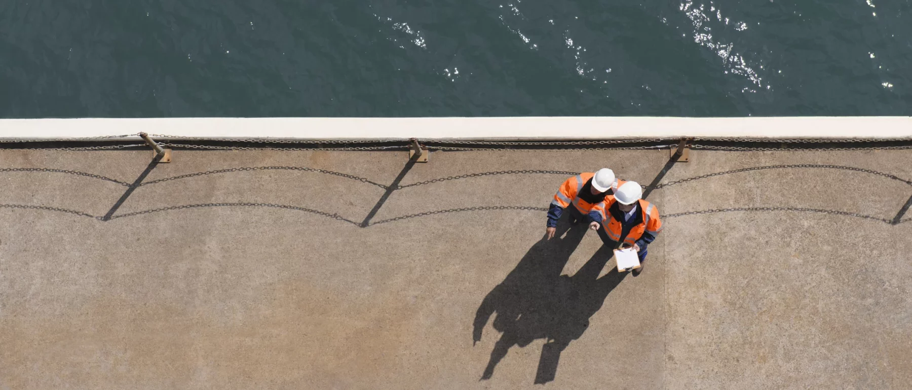 Top-down view of two workers in safety vests and helmets standing on a concrete dock next to a body of water, casting long shadows in the sunlight.