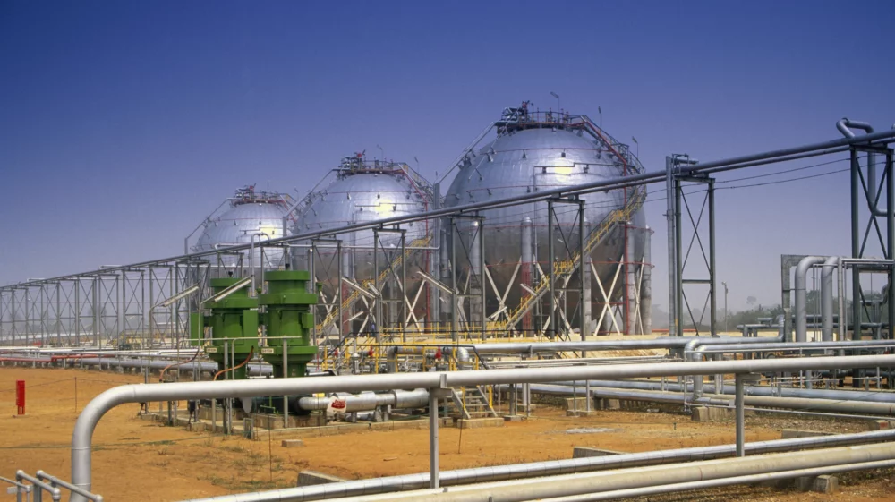 Three silver spherical oil tanks stand in a row behind a framework of metal pipes and a green pump.