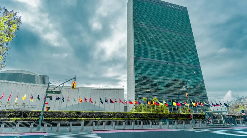 Exterior view of the United Nations headquarters in New York City, featuring the iconic tall glass building, surrounding structures, and rows of international flags lining the front.