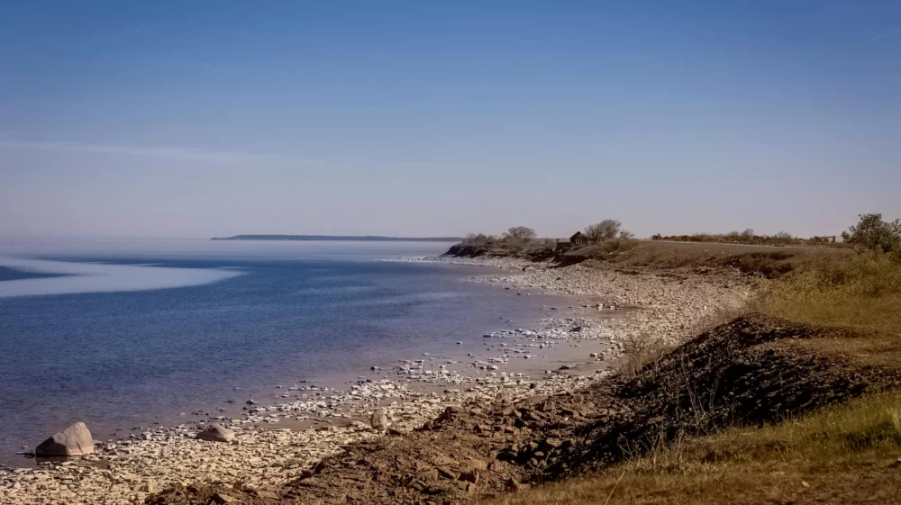 A serene coastline with calm blue waters stretching to the horizon, bordered by a rocky shore and grassy terrain under a clear blue sky.