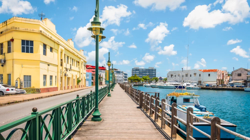 Bright image of wooden promenade at the waterfront of Bridgetown in Barbados. Colorful building against blue sky with white clouds. Boats and yachts in the harbor.
