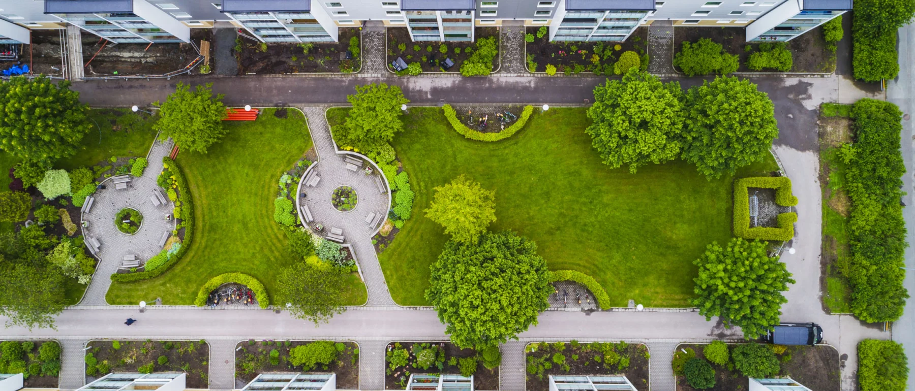 A view looking down on green grassy lawn with two gray gravel rings with benches, bushes, trees, bordered by sidewalk and two apartment blocks, horizontally framing the top and bottom of the image.