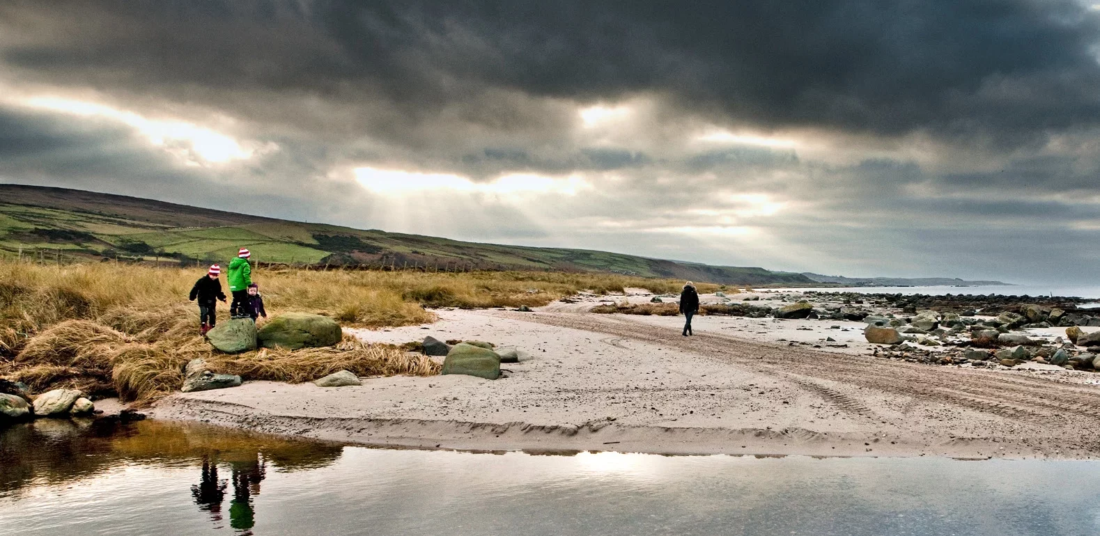 Gray cloudy skies above a beach, where water reflects the sky and three children perch on rocks between grass and sandy shore, and an adult walks away from the camera -- all in warm hats and parkas.