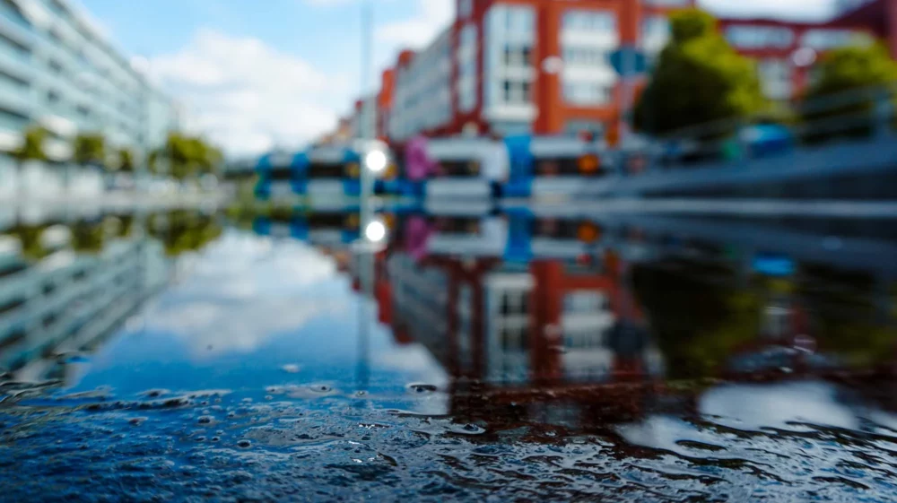 Blurry red and white buildings in the distance, with a water puddle in focus in the foreground reflecting blue sky and clouds.