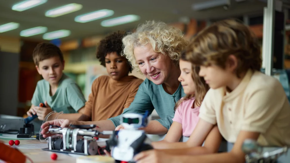 Smiling female adult assisting schoolchildren in making a futuristic robot.