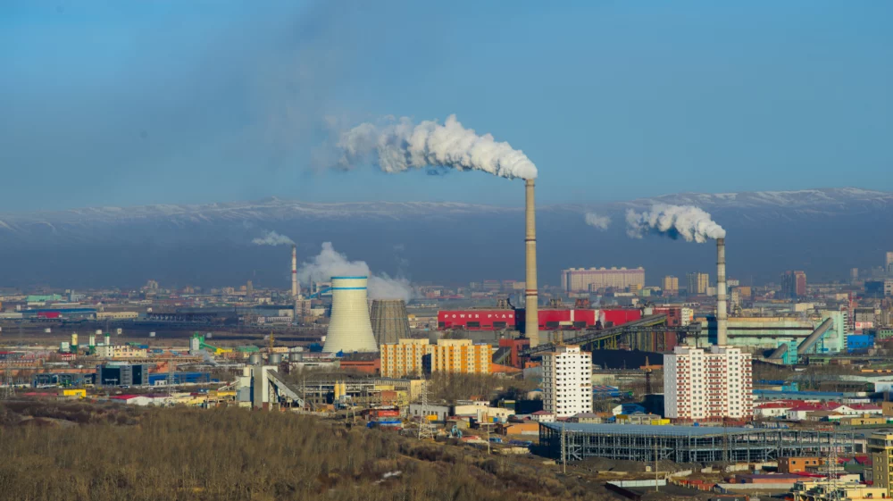 Elevated landscape with a power station in the foreground and the city of Ulaanbaatar behind. Snow-capped mountains sit under a blue sky in the background, behind a haze of smoke.