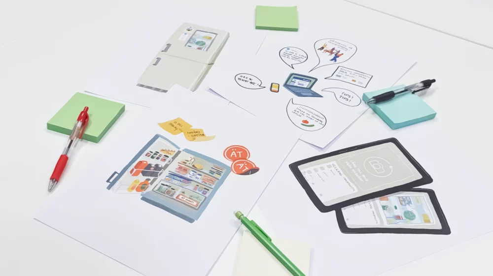Printed illustrations on a white desktop of a fridge organizing system, reminders on a closed fridge, phone app, website on a laptop screen, with pens and post-it notes scattered on top.