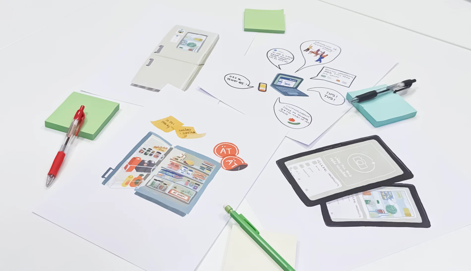Printed illustrations on a white desktop of a fridge organizing system, reminders on a closed fridge, phone app, website on a laptop screen, with pens and post-it notes scattered on top.