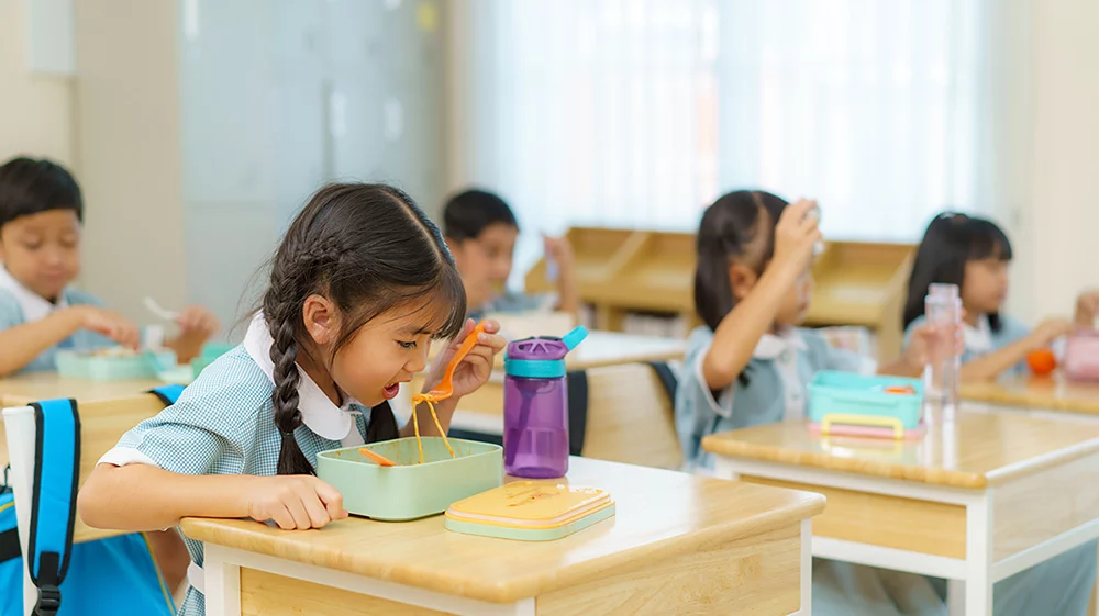 A schoolgirl eating noodles from a lunchbox at her desk, surrounded by classmates enjoying their meals in a bright classroom.