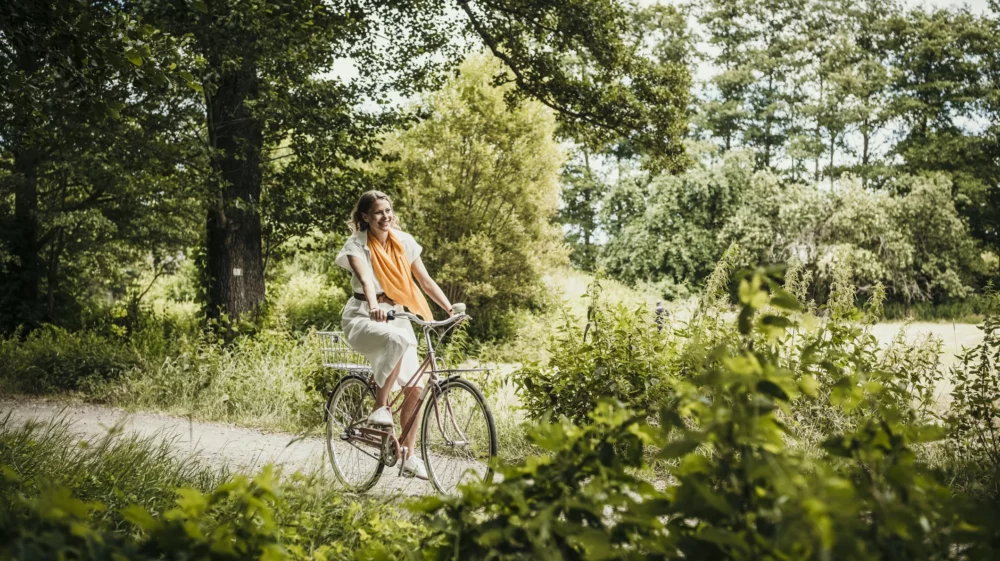 A happy woman riding a bicycle in the countryside road, surrounded by green trees, bushes and other plants.
