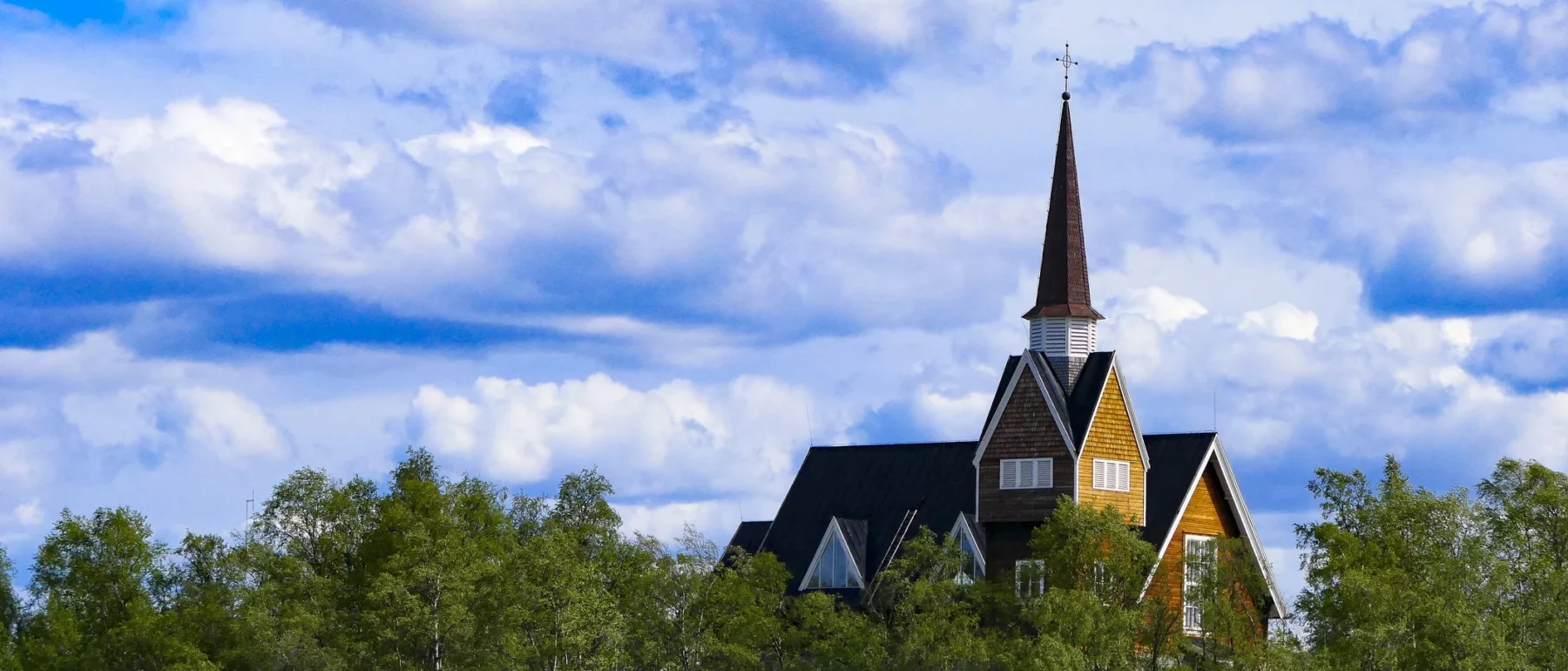 A wooden church with a pointed steeple in Karesuando, Sweden, surrounded by trees and water, under a blue sky with white clouds.