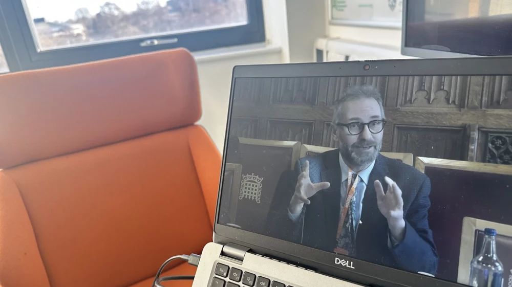 A laptop screen displaying SEI’s Kevin Hicks giving evidence at the UK House of Lords nitrogen inquiry. He is seated in a formal setting, gesturing with both hands while speaking. In the background, an orange chair and a window are visible in a well-lit office space.